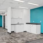 Modern office kitchenette with white cabinets, dark countertops, and a bold blue accent wall. The space has a grey tiled floor and fluorescent lighting.