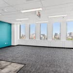 An empty office space with a blue accent wall, large windows offering a city view, carpeted floors, and fluorescent ceiling lights.
