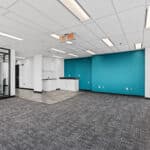 Modern office interior with teal accent walls, carpeted floors, overhead lighting, white cabinetry, glass doors, and an exit sign.