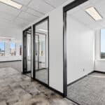 Modern office space with glass partitions, carpeted and tiled floors, and a city view through large windows under a bright, artificial light.